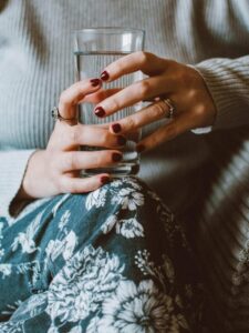 The lower part of the body of a woman sitting on a stool wearing a flower pattern dress and holding a glass of water in her hand.